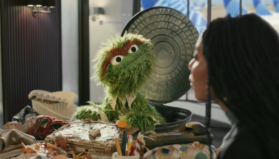 Oscar the Grouch in a trash can, wearing a tie and shirt collar. In a conference room with a woman. The table is covered in trash.