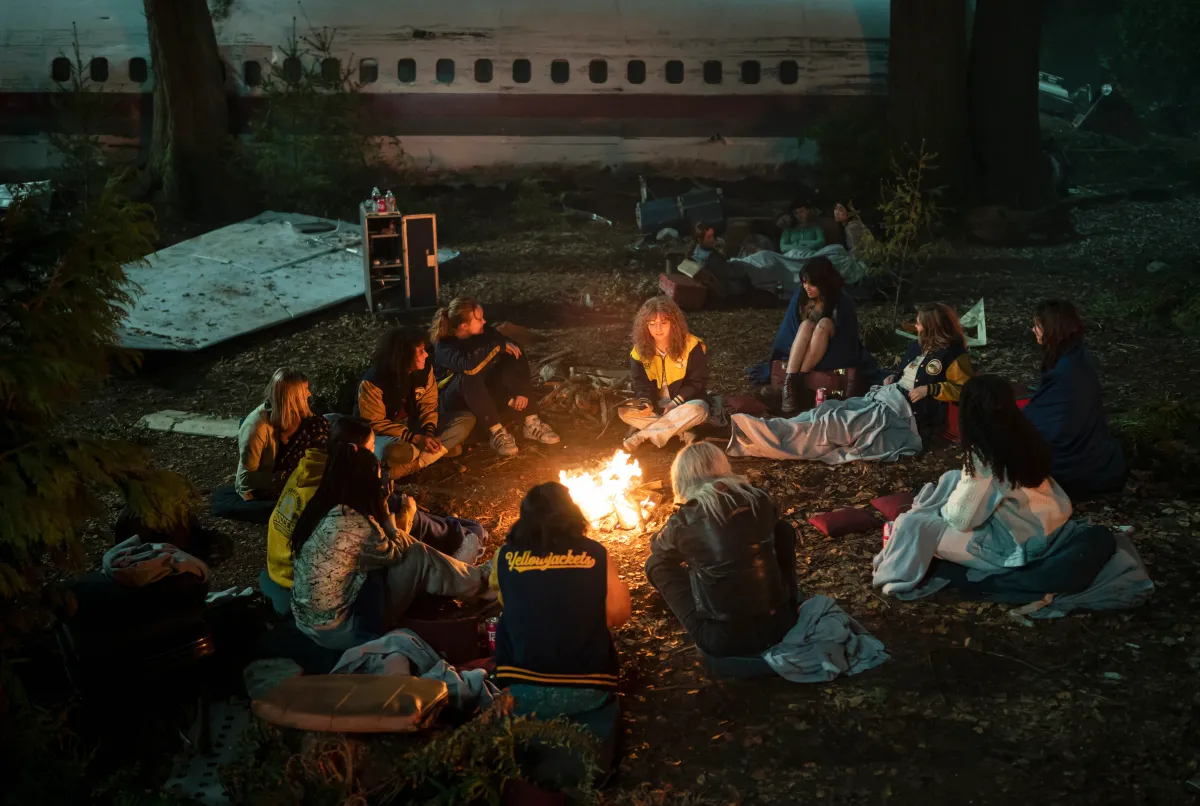 A group of teenage girls sit in a circle around a campfire next to a crashed plane, in a still from YELLOWJACKETS