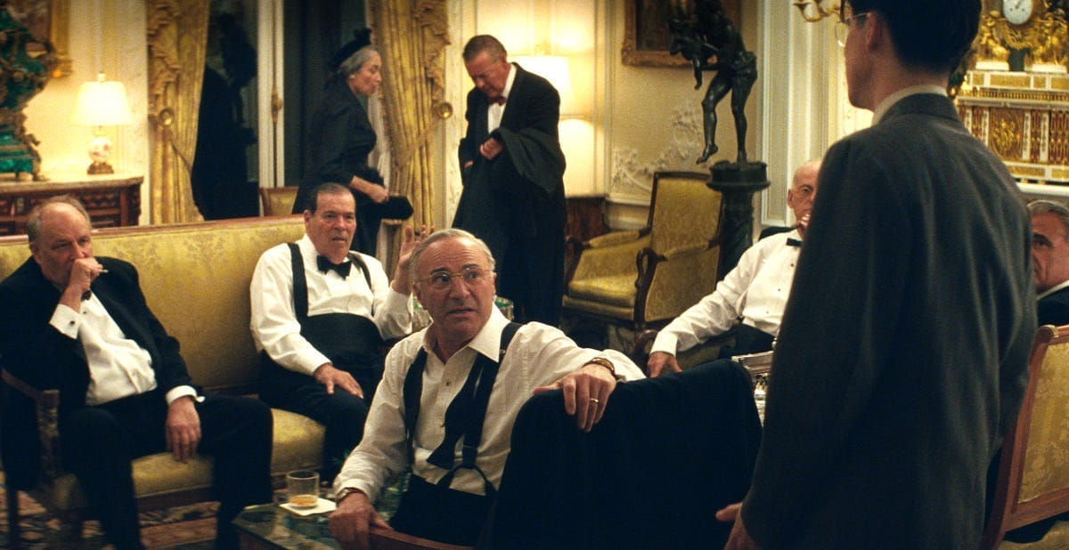 Timothee Chalamet stands in front of a room full of wealthy men in a scene from Marty Supreme