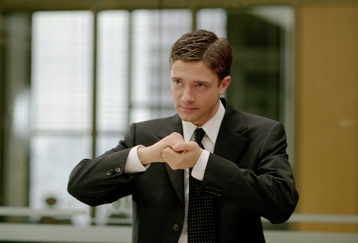 Screencap from In Good Company: Topher Grace in a suit, clasping his hands together to symbolize synergy.