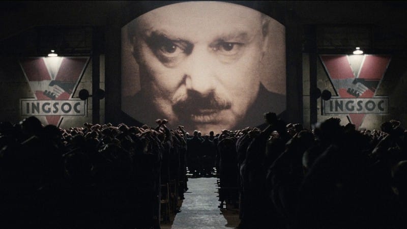 Screencap 1984: A giant image of Big Brother looks down on an assembled crowd. Large INGSOC banners hang on either side of the monitor.