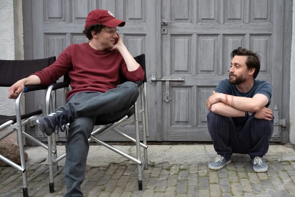 Jesse Eisenberg sits in a folding chair staring at Kieran Culkin, who squats next to him