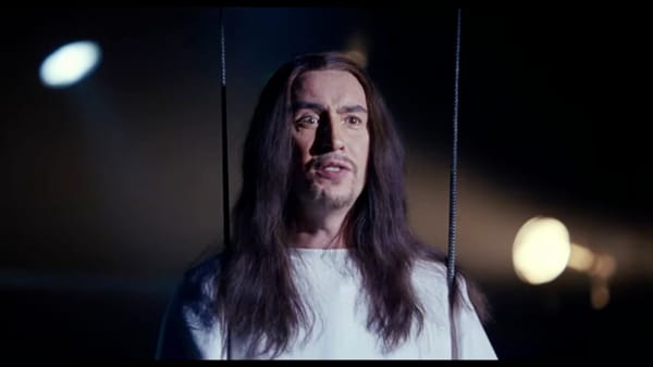 Steve Coogan with long hair and a white robe suspended from wires above a high school theater stage (playing Jesus).