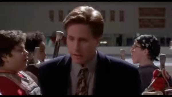 Gordon Bombay in a suit and tie at the hockey rink, walking between two rows of Mighty Ducks.