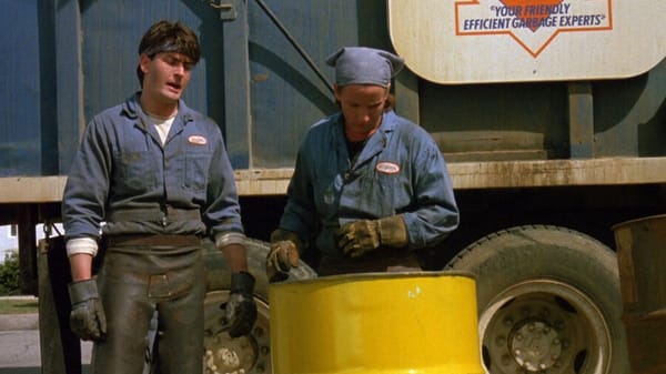 Charlie Sheen and Emilio Estevez dressed as garbage men. They're looking into a yellow barrel. Source: Men at Work