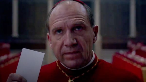 Ralph Fiennes dressed as a Catholic cardinal holds up his ballot to cast a vote for the next pope.