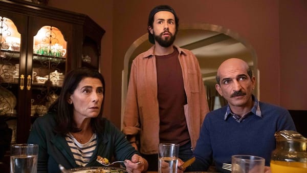 Two parents sit eating while their adult son stands over them in a scene from Ramy