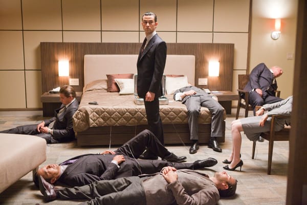 A man stands in a hotel room full of well-dressed people sleeping in a scene from Inception